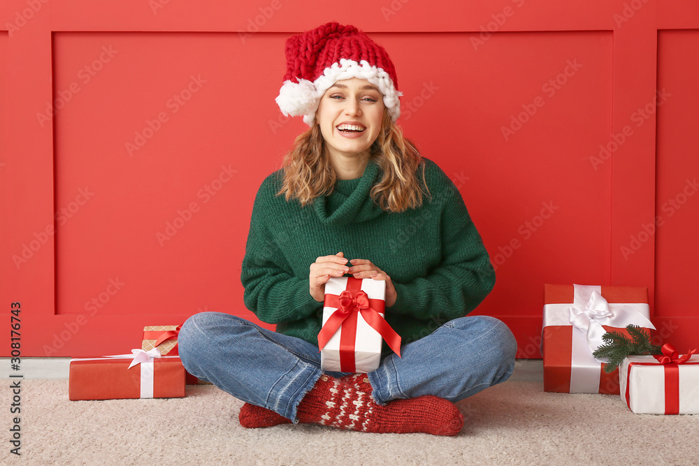 Happy young woman in winter clothes and with Christmas gifts sitting near color wall