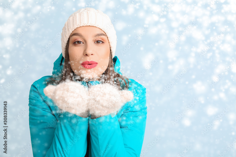 Happy young woman in winter clothes blowing snow on color background