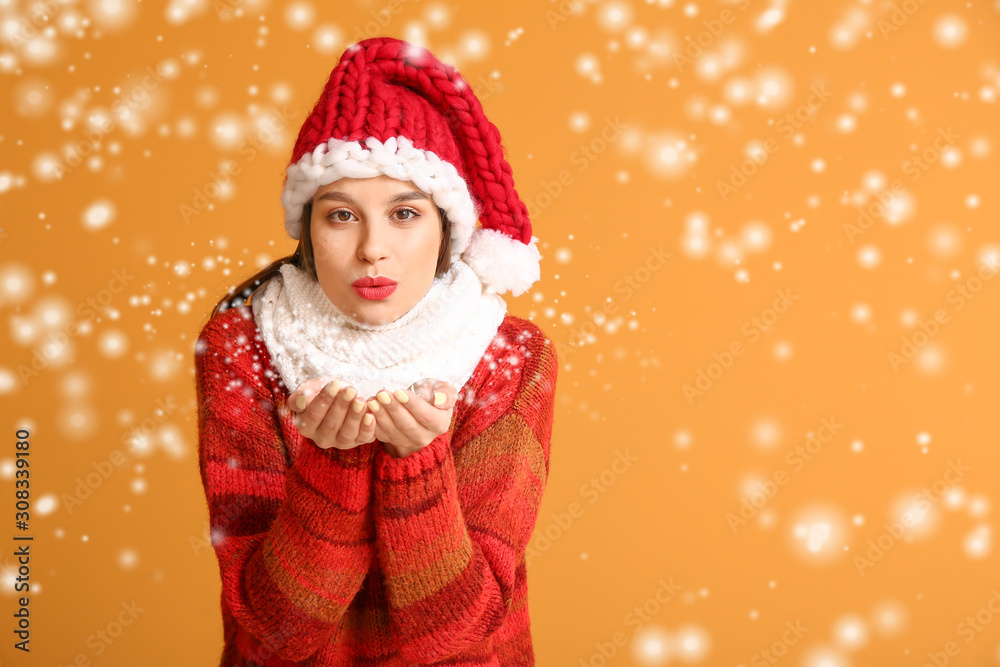 Young woman in winter clothes blowing snow on color background