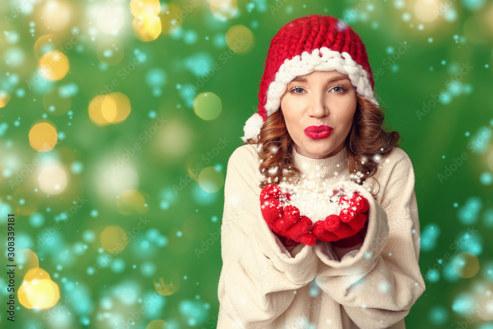 Happy young woman in winter clothes blowing snow on color background