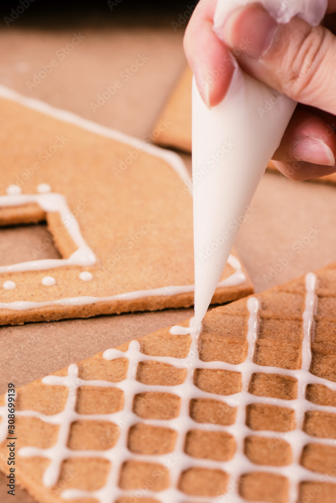 Woman is decorating gingerbread cookies house with white frosting icing cream topping on wooden tabl