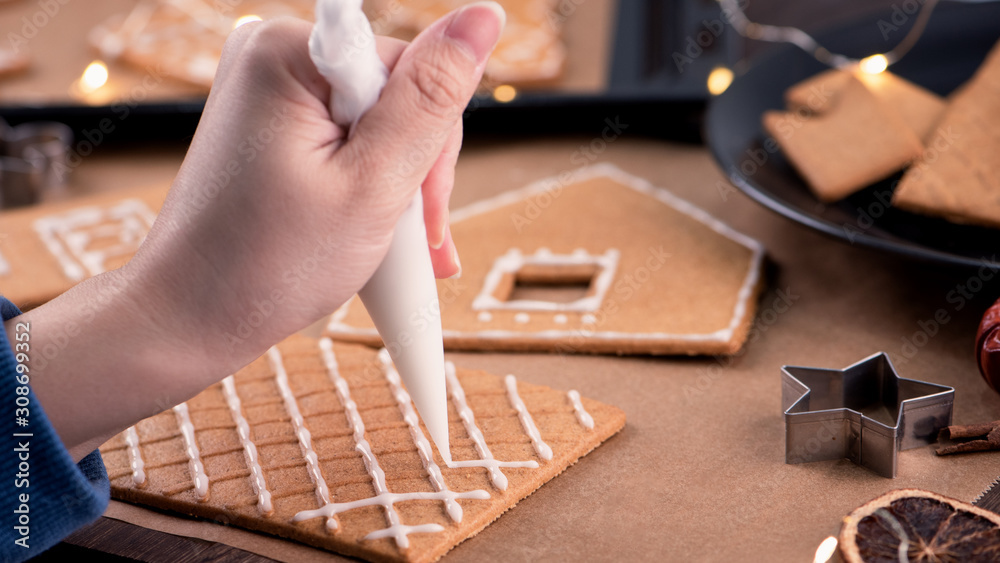 Woman is decorating gingerbread cookies house with white frosting icing cream topping on wooden tabl