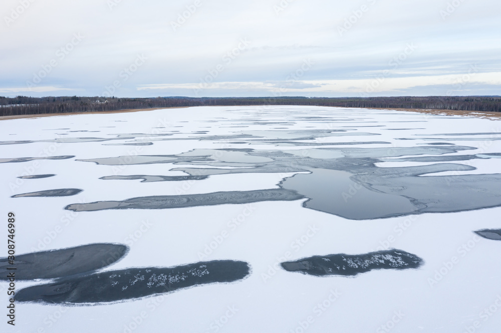 Aerial drone view of a white icy frozen lake with abstract unfrozen water patches in sunrise light. 