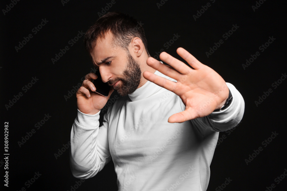 Portrait of young man with mobile phone showing stop gesture on dark background