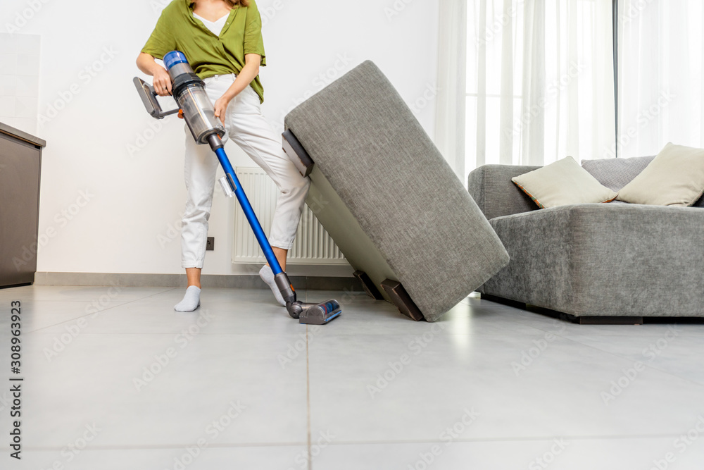 Young woman cleaning floor under the sofa with cordless vacuum cleaner at home. Concept of easy clea