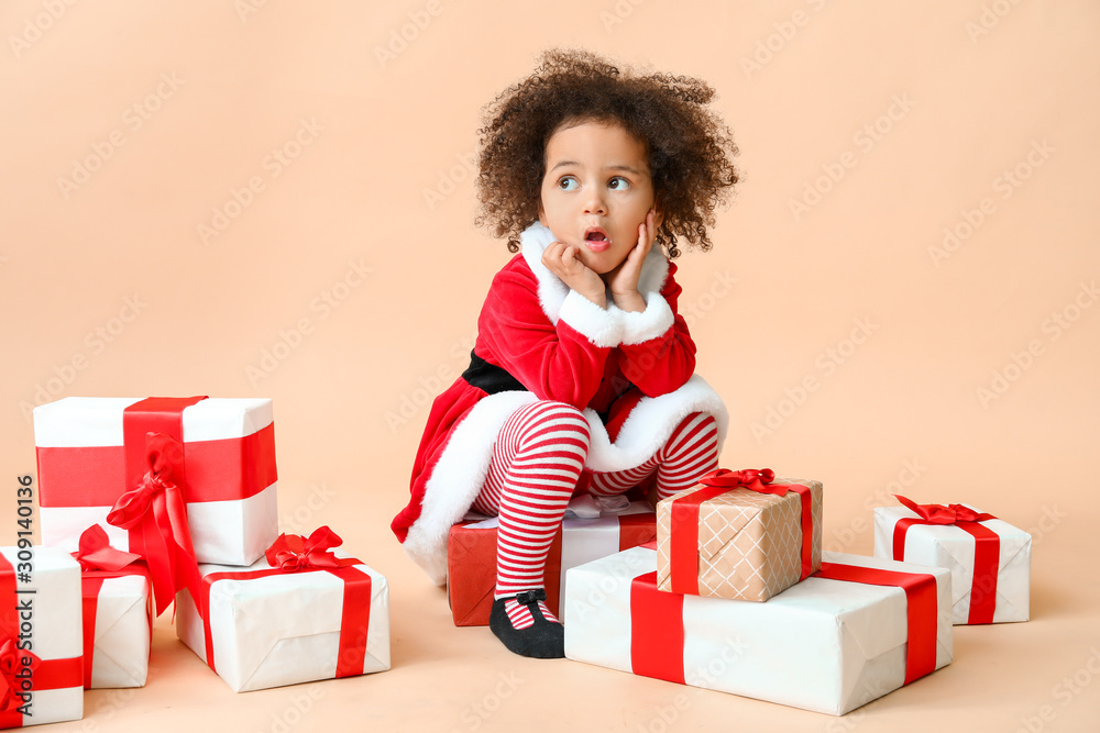 Little African-American girl in Santa costume and with gift boxes on color background