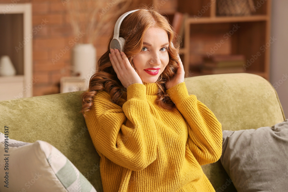 Happy young woman in winter clothes listening to music on sofa at home