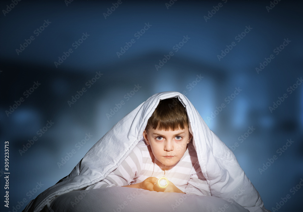 Serious child with flashlight hiding under blanket