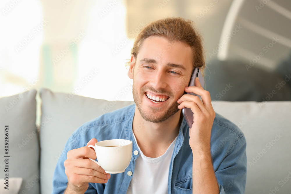 Handsome man with cup of coffee talking by mobile phone at home