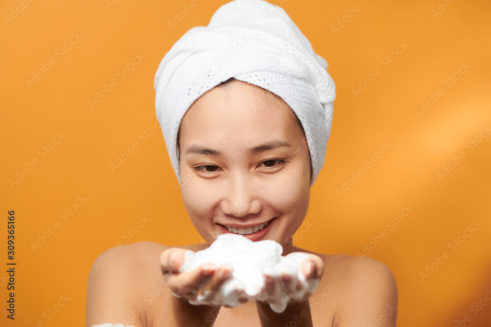 Funny Asian woman in a towel on the head happy cleanses the skin with. Isolated on orange background