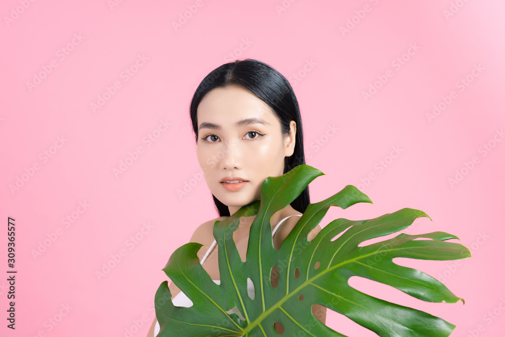 Portrait of young beautiful Asian woman holding tropical leaf on pink background. Natural beauty con