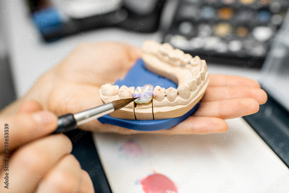 Dental technician coloring dental prosthesis with a paint brush at the laboratory, close-up view. Co