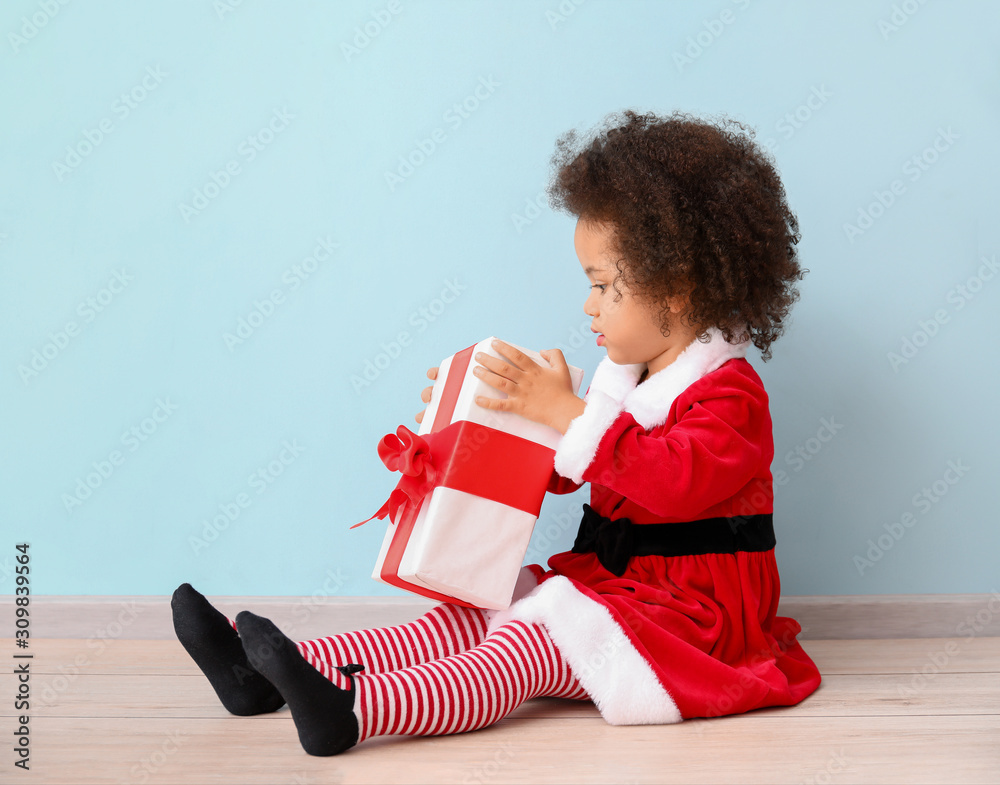 Little African-American girl in Santa costume and with gift near color wall