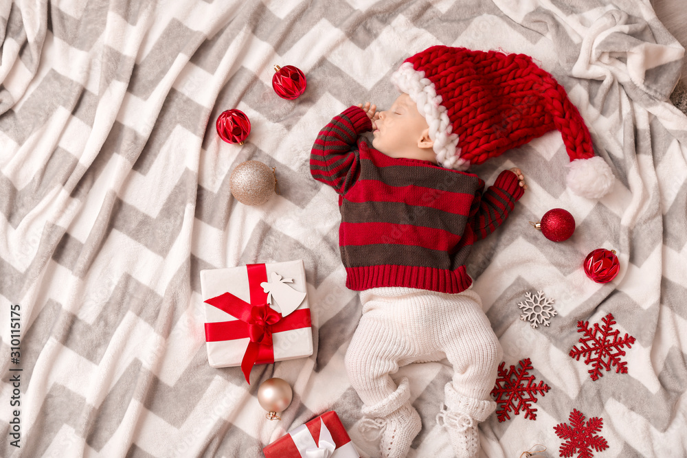 Cute little baby in Santa Claus hat and with Christmas gifts sleeping on plaid, top view