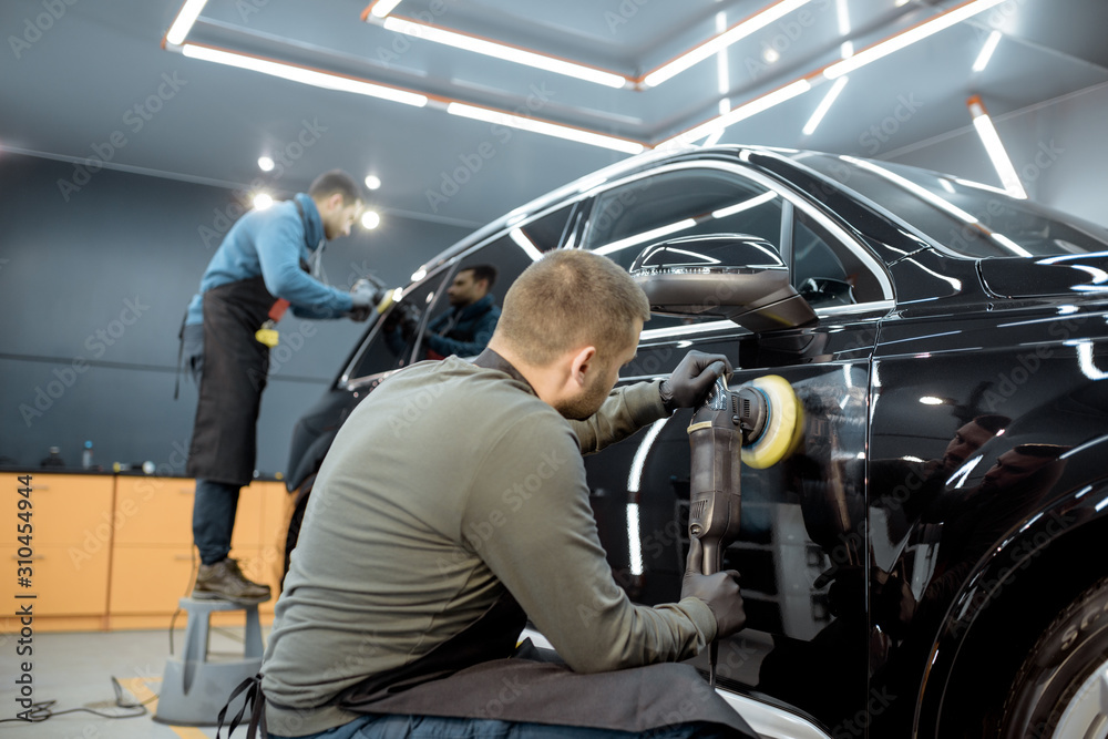 Two workers polishing vehicle body with special grinder and wax from scratches at the car service st