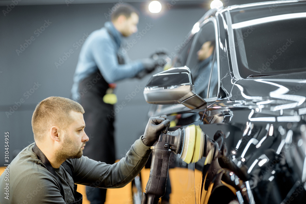 Two workers polishing vehicle body with special grinder and wax from scratches at the car service st