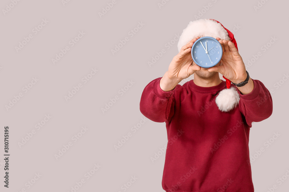 Young man in Santa hat and with alarm clock on light background. Christmas countdown concept