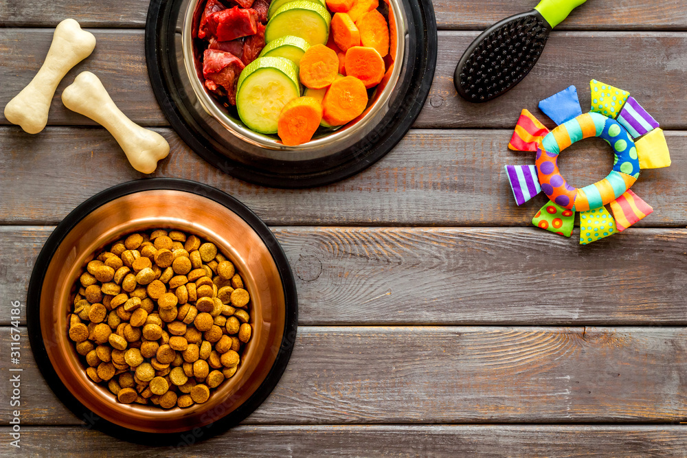 Dog feed - dry food and raw meat - near toy, brush and cheving bones ondark wooden background top vi