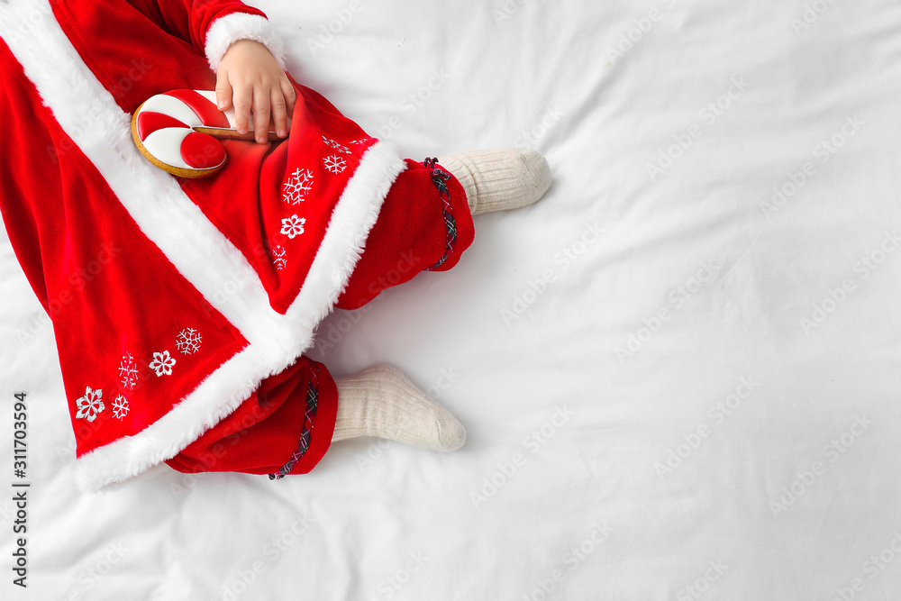 Cute little baby in Santa Claus costume and with cookie lying on bed, top view