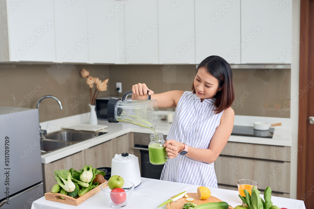 年轻的亚洲女人在现代厨房里把绿色奶昔倒进玻璃里