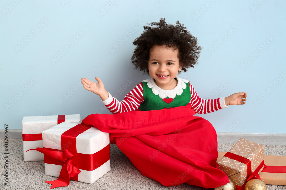 Little girl in costume of elf, with Santa Claus bag and Christmas gifts sitting near color wall