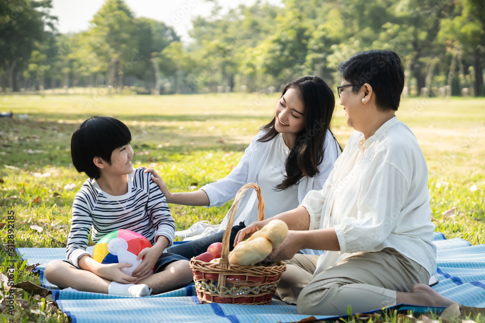 亚洲家庭一起在清新的绿色公园野餐。妈妈笑着和孩子说话，奶奶