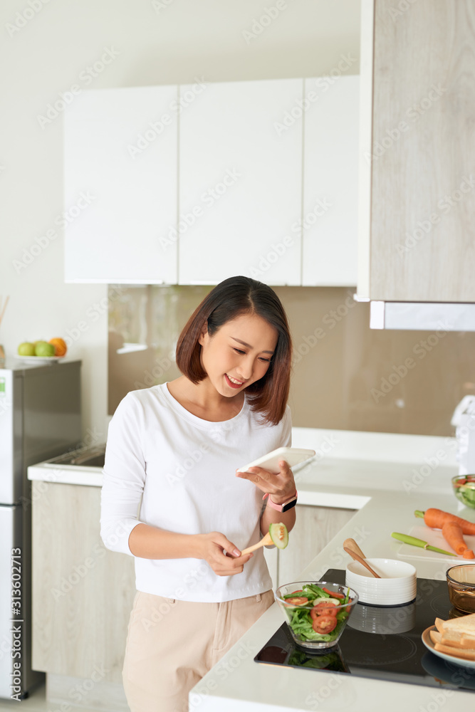 Beautiful young woman wearing apron cooking healthy salad at the kitchen at home, using mobile phone