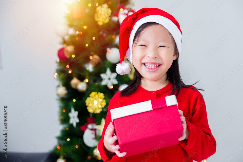 Little asian girl wearing santa claus hat and holding gift box with smiles in front of christmas tre