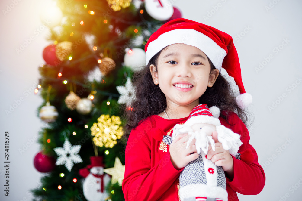 Little asian girl wearing santa claus hat and holding doll with smiles in front of christmas tree.