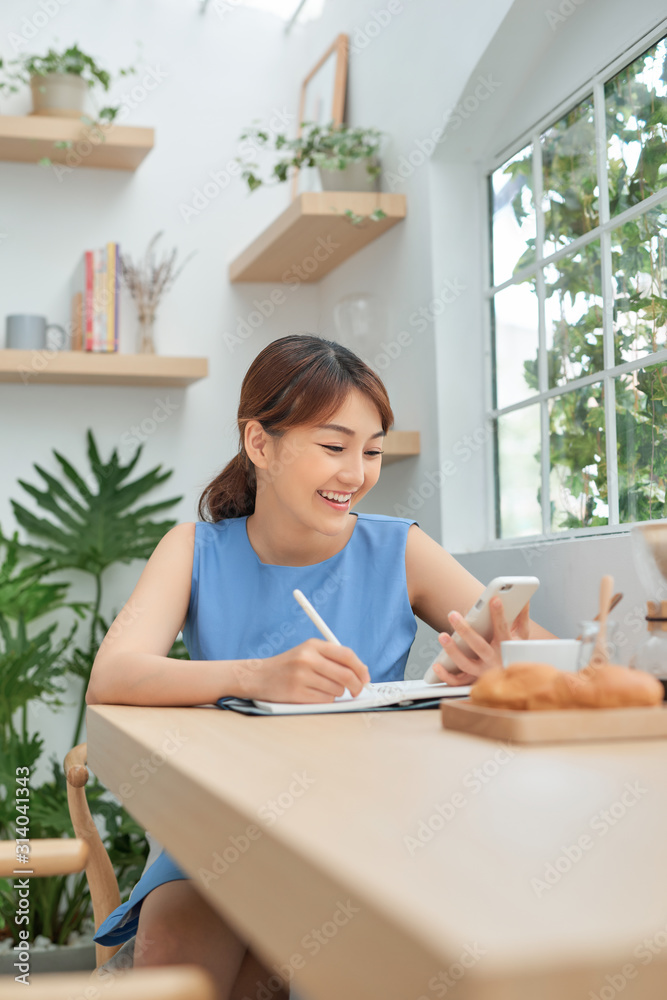 Smiling young Asian female entrepreneur sitting at a table and writing down notes