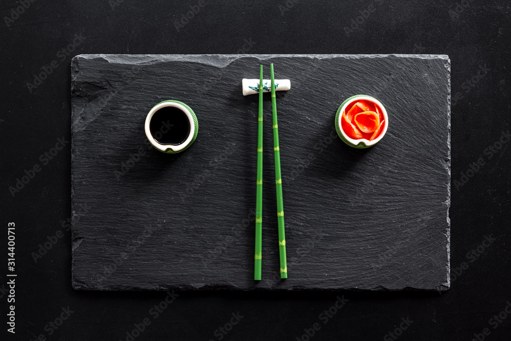 Table served for eating sushi. Chopsticks, small bowls with ginger and sause, mat on black backgroun