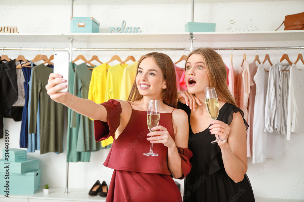 Beautiful young women drinking champagne and taking selfie in shop with modern clothes