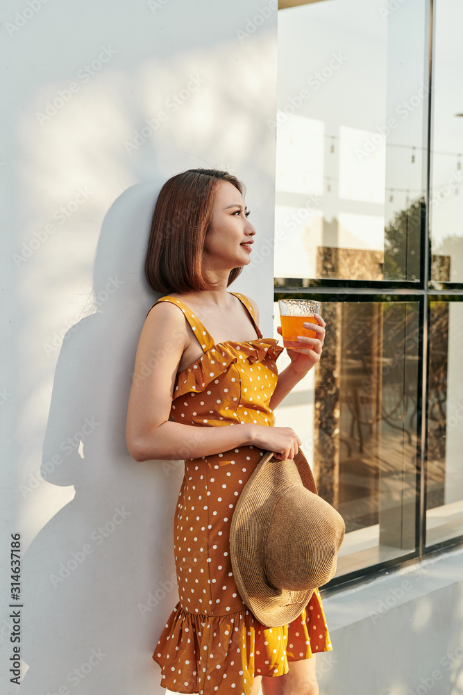 漂亮的亚洲女孩穿着复古连衣裙，手里拿着一顶墙后有橙汁的帽子