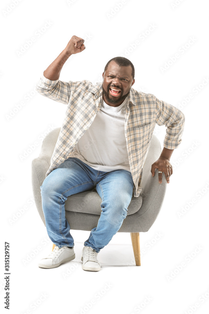 Emotional African-American man watching TV while sitting in armchair against white background