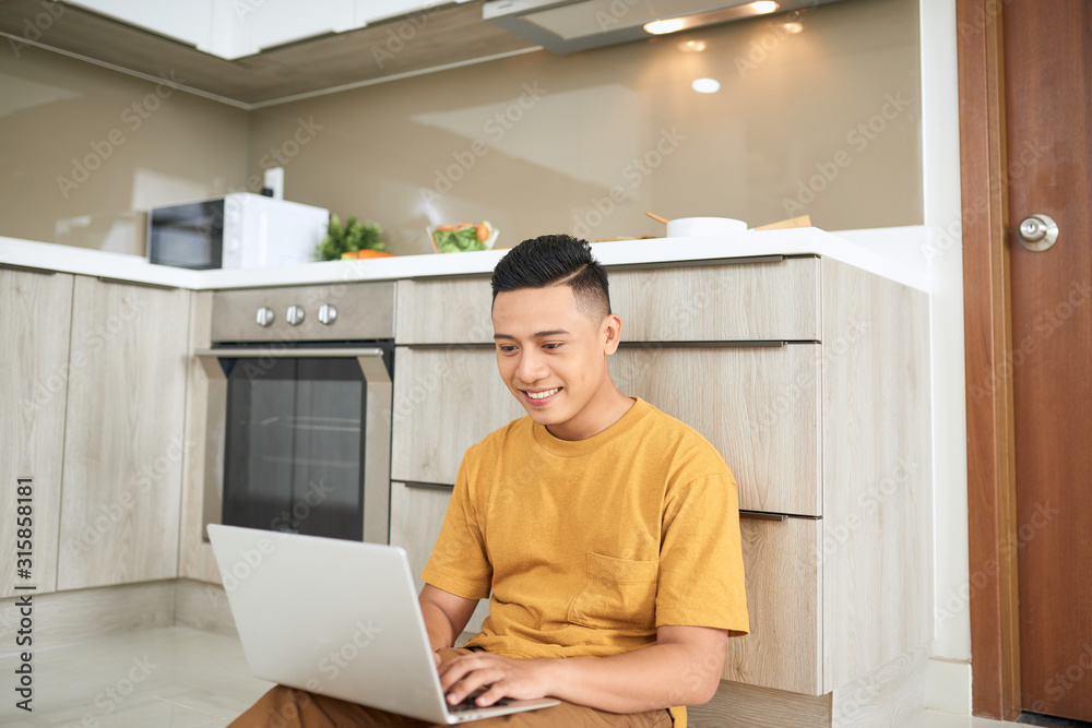 attentive asian man using laptop while sitting on floor in kitchen