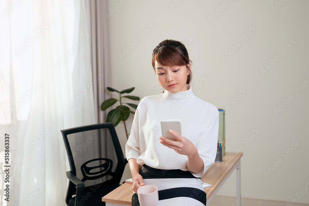 Smiling young asian businesswoman using mobile phone while drinking coffee at the office