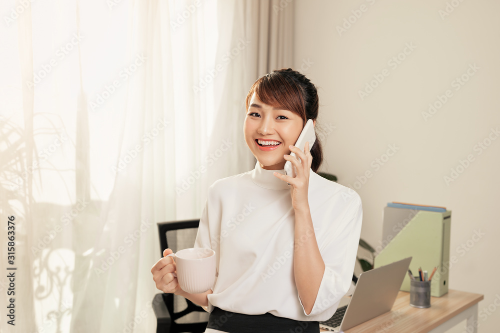 Young Asian businesswoman talking on phone while drinking coffee at the table in office