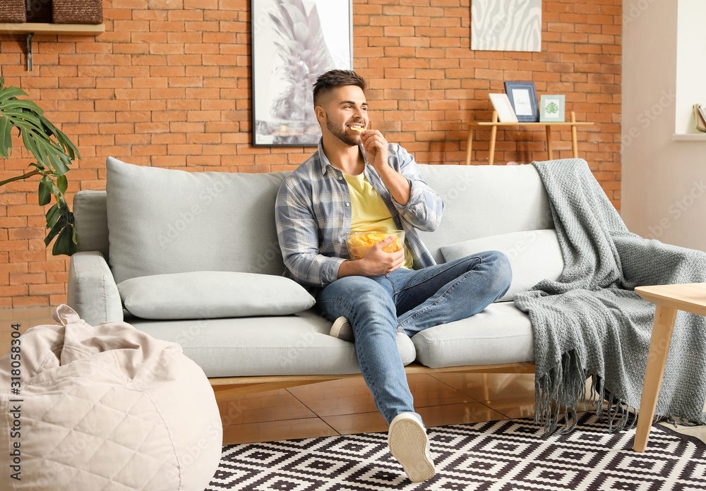 Handsome young man eating tasty potato chips at home