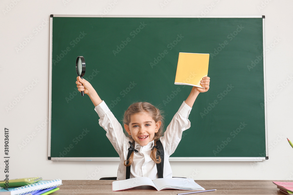 Happy little schoolgirl sitting at desk in classroom