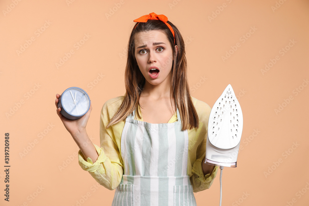 Stressed young housewife with iron and alarm clock on color background