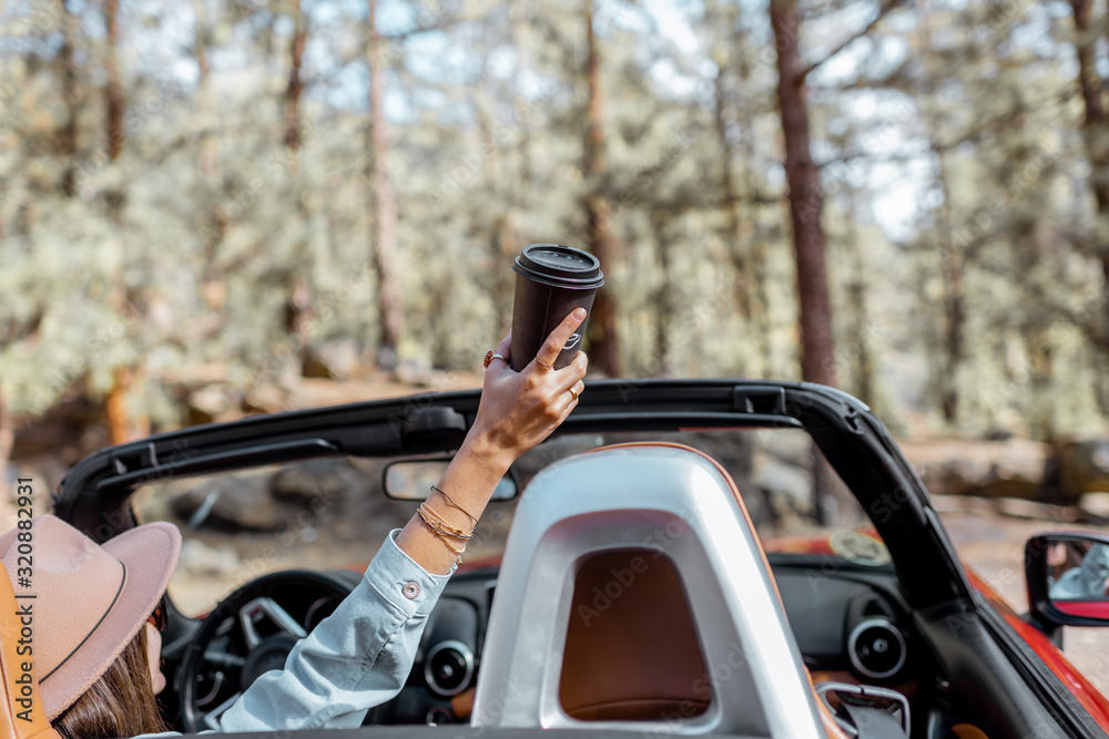 Woman enjoying traveling by a cabriolet in the forest, raising hands up with a coffee cup, view from