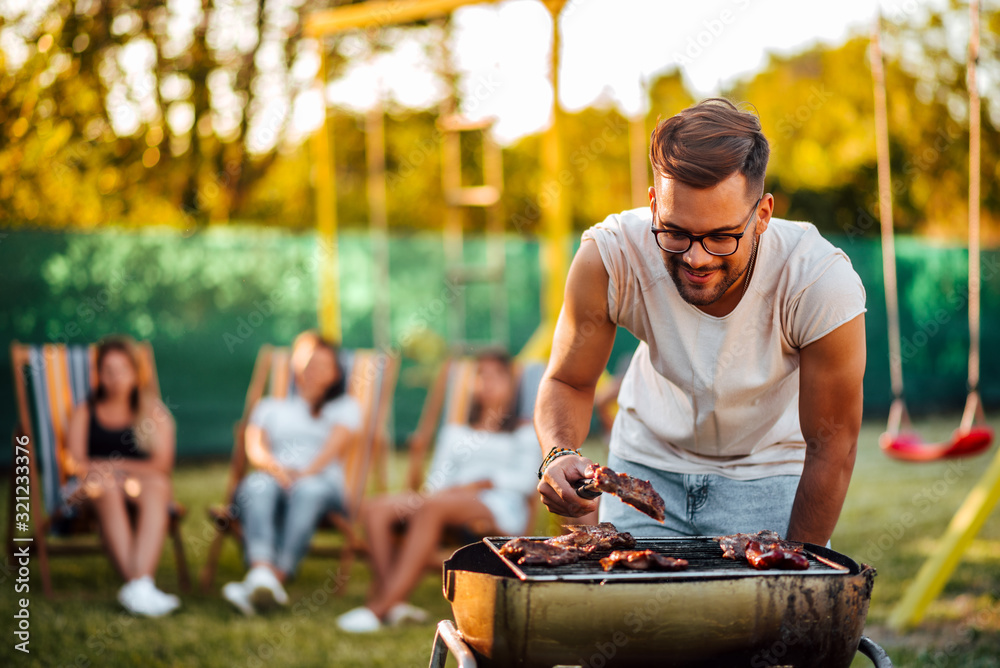 Young positive man grilling meat on a barbecue outdoors, for friends.