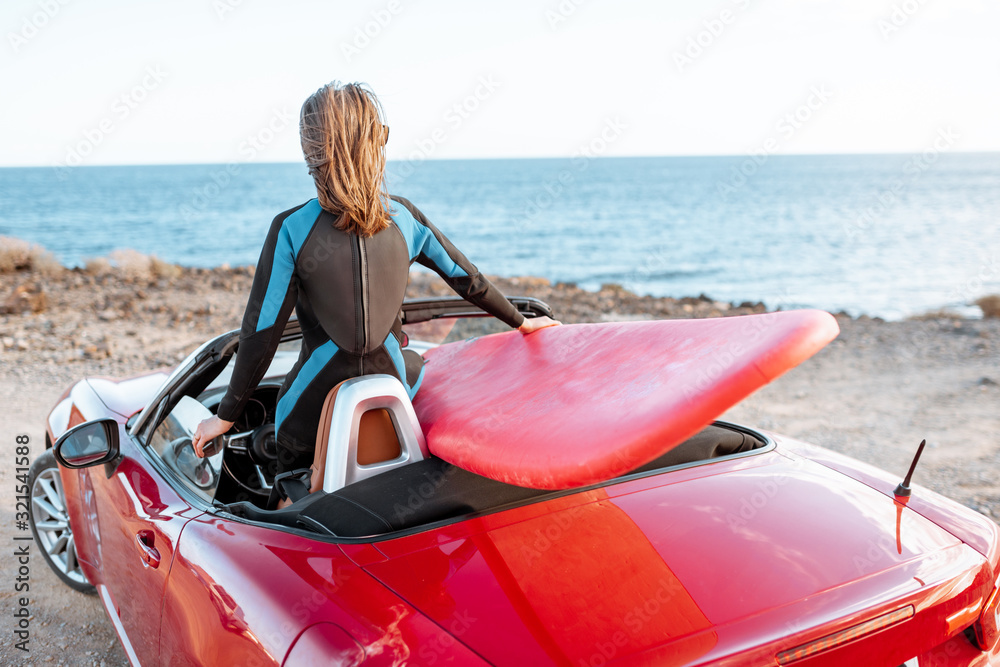 年轻的女冲浪者在岩石海岸驾驶带冲浪板的红色敞篷车。无忧无虑的生活方式和