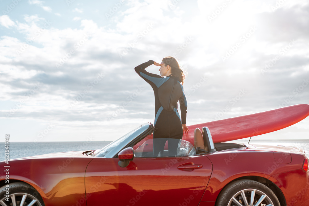 Woman enjoying beautiful landscapes while standing on her sports car with a surfboard near the ocean