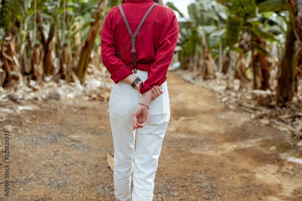 Woman as a tourist or farmer dressed casually in red and white walking between banana rows at the pl