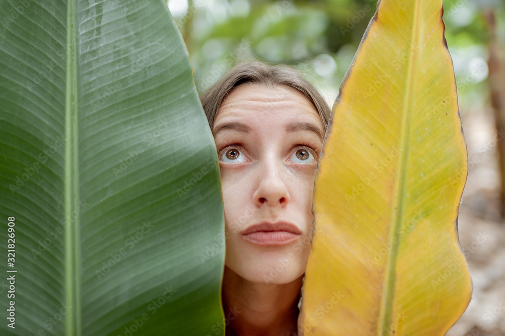 Facial portrait of a woman with sad emotions hiding behind a fresh and dry banana leaves on the plan