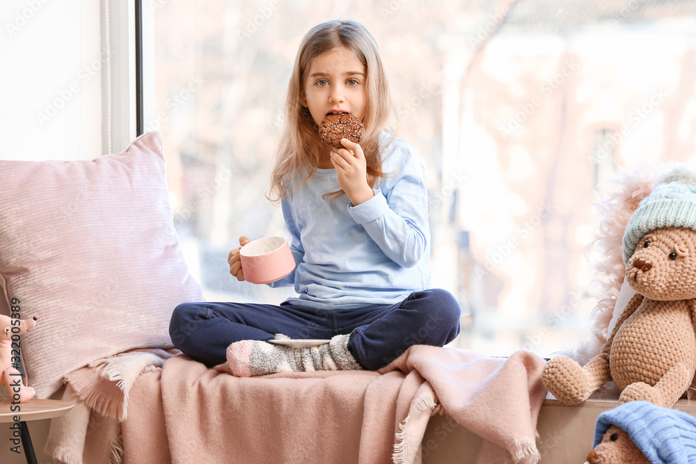 Cute little girl drinking hot chocolate with cookie on window sill at home