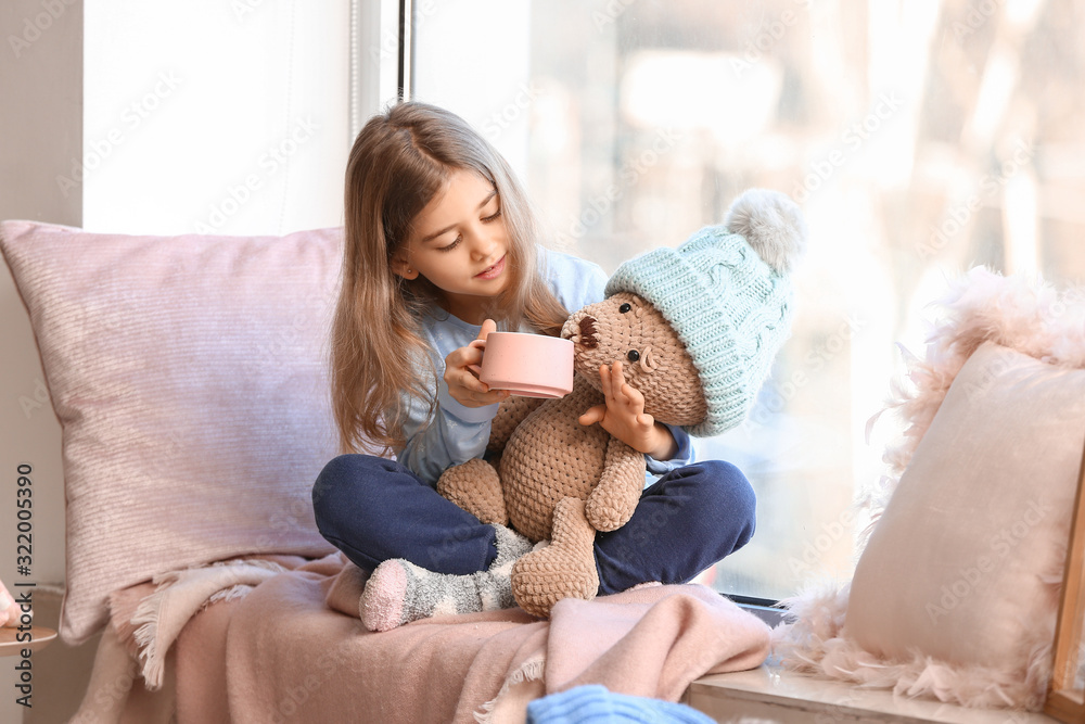 Cute little girl with teddy bear drinking hot chocolate on window sill at home