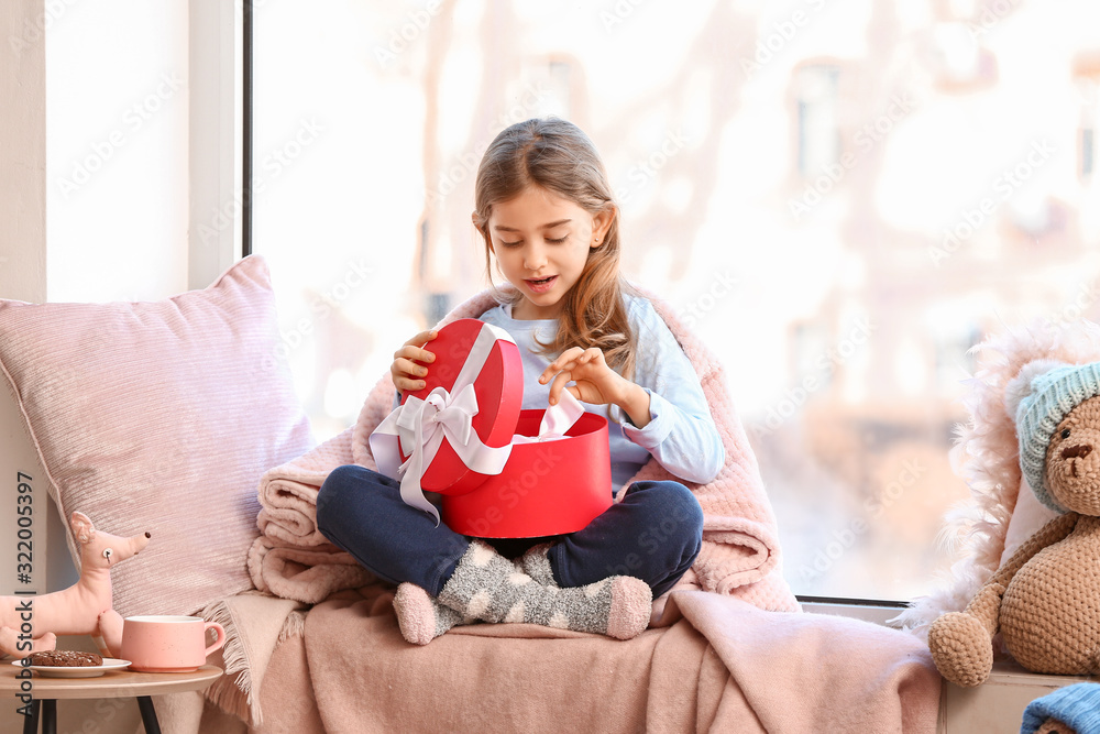 Cute little girl opening gift box on window sill at home