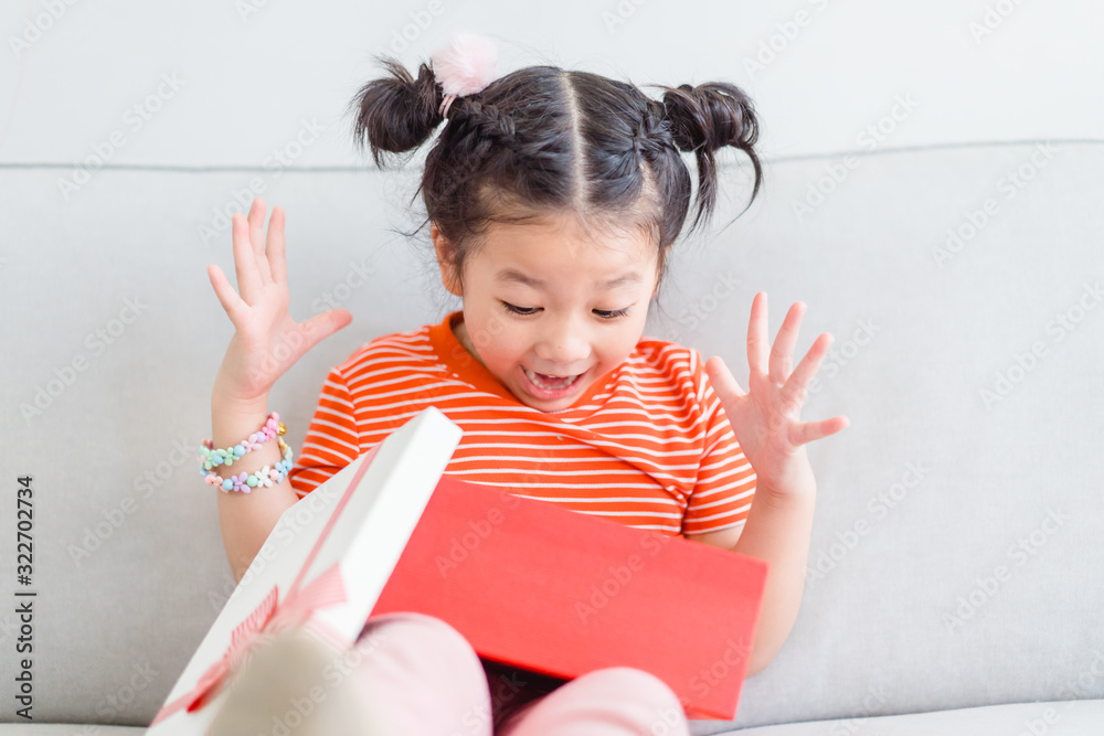 Little asian girl smile and excited and holding red gift box on sofa in living room background.child